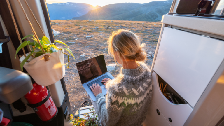 A woman enjoys working remotely out of an RV in front of a beautiful mountainscape