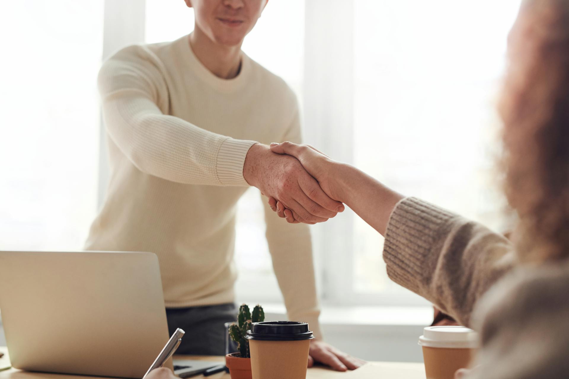 Two business professionals shaking hands across a desk in a bright office, symbolizing executive hiring and partnership.