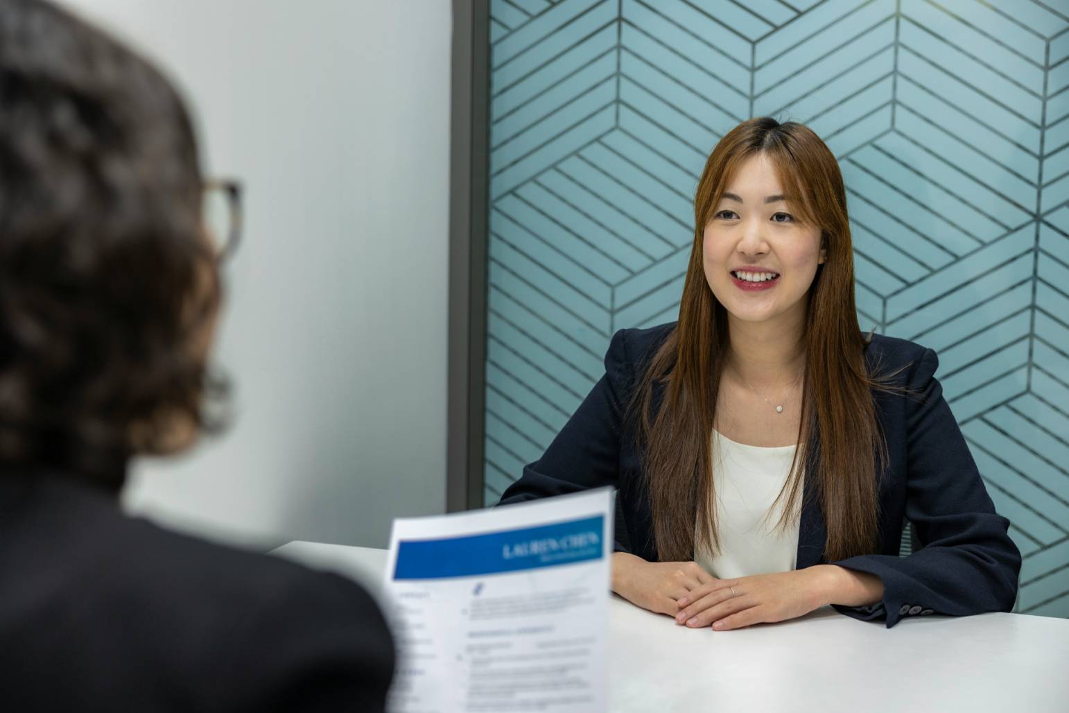 A businesswoman in a blazer sits at a table during a job interview, facing an interviewer who is holding her resume. 